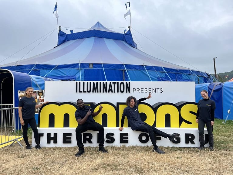 Two people dressed in black lounging on a 'Minions' hoarding in front of a blue event tent.